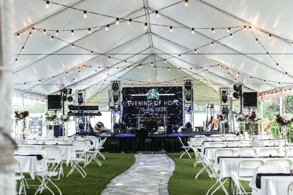 Evening of Hope Event - tent on the lawn overlooking Lemon Bay with strings of lights, a band platform, tables, chairs, and floral centerpieces